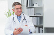 © WavebreakMediaMicro - Senior male doctor in white coat sitting at clinic desk holding clipboard with stethoscope draping