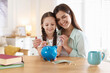 © New Africa - Mother and daughter putting pocket money into piggybank at table indoors
