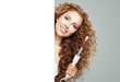 © artmim - Cute young model woman with long curly wavy hairstyle showing hot curling tongs iron and empty paper banner placard on white background