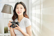 © Dialogue Frame - Young Asian Woman Photographer in White T-Shirt Holding Vintage Camera by Bright Window Indoors