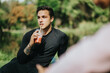© qunica.com - A young man enjoying a smoothie in a natural outdoor park setting, wearing casual attire, creating a feeling of leisure and enjoying a sunny day.