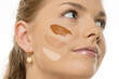 © vladimirfloyd - Close-up portrait of smiling young woman with different foundation shades swatched on her cheek, demonstrating makeup color matching process against white background