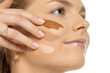 © vladimirfloyd - Close-up of young woman with contemplative expression displaying three different foundation shades swatched on cheek, comparing makeup colors against white background.