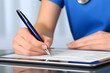 © Health Studio - A person in blue scrubs writes on a digital tablet with a pen, possibly filling out medical or professional documents.