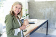 © Wavebreak Media - Mature woman standing at wooden desk in office holding mug by smartphone, documents, plant, smiling