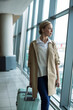 © Wavebreak Media - Woman wearing trench coat standing in airport hall holding phone rolling blue suitcase, copy space