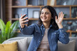 © Tetiana - Smiling young Indian woman sitting on sofa at home, talking on video call and greeting on mobile phone camera
