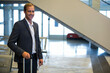 © Wavebreak Media - Smiling man wearing business attire, holding suitcase at modern airport with escalator, copy space