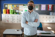 © Wavebreak Media - Teacher wearing black mask standing in classroom with bulletin board and desks with notebook