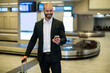 © Wavebreak Media - Mid adult man wearing black blazer checking smartphone and holding suitcase at airport carousel