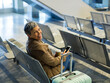 © Wavebreak Media - Senior woman sitting airport chair beside mint green suitcase holding coffee cup, phone, copy space