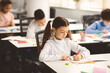 © Prostock-studio - Cute female pupil sitting at desk in classroom at the elementary school. Student girl doing test in primary school. Children writing notes during the lesson or drawing picture in diary
