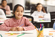 © Prostock-studio - Elementary Education. Portrait of small smiling asian girl sitting at table in classroom at primary school or kindergarten with group of classmates. Return back to school after coronavirus quarantine