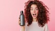 © sablengjago - Amazed woman with curly hair holding a dark grey bottle against pink background.