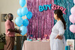 © pressmaster - Pregnant Asian woman standing with hand on hip facing Black man holding blue balloons at gender reveal party