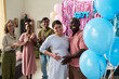 © pressmaster - Pregnant Asian woman and Black man standing together smiling while friends clapping and colorful balloons decorating background