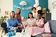 © pressmaster - Happy young pregnant Caucasian woman holding belly surrounded by friends celebrating event during gender reveal party at home