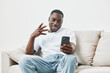 © SHOTPRIME STUDIO - Smiling African American man using smartphone at home in casual clothing, enjoying video call, relaxed atmosphere and cheerful vibe in bright living space