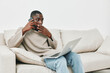 © SHOTPRIME STUDIO - Young Black man in casual sweater talking on the phone while using a laptop at home, expressing concern and focus during a conversation