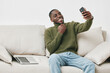 © SHOTPRIME STUDIO - Smiling African American man in green sweater taking selfie on smartphone, relaxed on couch with laptop, modern home interior, cheerful and casual atmosphere
