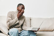 © SHOTPRIME STUDIO - A surprised young Black man in a cozy sweater sitting on a sofa, using a laptop, expressing disbelief or shock, showcasing emotions and casual lifestyle
