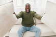 © SHOTPRIME STUDIO - Happy young Black man expressing joy while sitting on a sofa, holding decorative pillows in a cozy living room setting with soft lighting