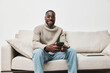 © SHOTPRIME STUDIO - Happy young man in a cozy sweater using a smartphone on a neutral sofa, conveying comfort and modern lifestyle