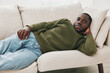 © SHOTPRIME STUDIO - Relaxed young African man in a green sweater lying on a beige couch with a peaceful expression, showcasing comfort and casual lifestyle at home