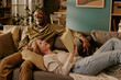 © AnnaStills - Senior Caucasian woman lying on sofa holding book smiling up at senior Black man sitting beside her laughing, couple relaxing together in living room showing romantic connection