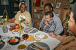 © Seventyfour - Smiling family with little Black boy drinking from glass sitting at dining table enjoying conversation over festive dinner at home, camera flash