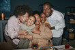 © Seventyfour - Portrait of smiling Black family members embracing and enjoying quality time at dining table during festive gathering at home, camera flash