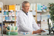 © JackF - Confident professional senior pharmacist in white coat working behind drugstore counter, inputting purchase orders into computer system, managing incoming supplies and ensuring stock availability