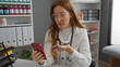 © Krakenimages.com - Woman doctor in clinic interior, redhead and attractive, checks smartphone while holding pen, surrounded by medical supplies, in a professional indoor setting.