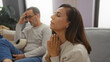 © Krakenimages.com - Middle-aged hispanic couple indoors with woman praying and man in background, showing love and family dynamics in cozy living room setting.
