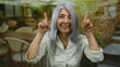 © Krakenimages.com - Senior woman with grey hair pointing upwards on an outdoor restaurant terrace with a street view, embodying vibrant energy.