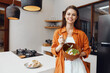 © SHOTPRIME STUDIO - Young woman smiling while holding a salad bowl in a modern kitchen, showcasing healthy eating and vibrant lifestyle choices for seasonal living