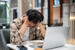© bongkarn - Man in plaid shirt holding hair thinking or stressing in front of laptop and notebook at cafe table.