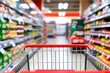 © sujgat - Part of shopping trolley in supermarket aisle. Blurred shelves in grocery store and trolley. Copy space for text, shopping cart handle with yellow grip red accents, Shopping cart in supermarket.