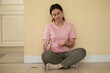 © Yuliia - woman sits on the floor against a wall, holding a glass of water and pills, looking tired and in discomfort.