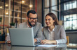 © 44designstore - Two smiling professional business man and woman, sitting together at a desk in a modern industrial-style office, collaborating and looking at a laptop screen