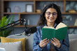 © Tetiana - Close-up photo of a young Indian girl at home, sitting on the couch and reading into a microphone from a book she is holding