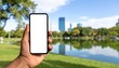 © mudrika - A person holding a blank smartphone in a park by a serene lake with city skyline in the background