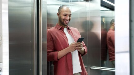 Smiling businessman is using mobile phone while standing in the elevator in modern office building. Happy worker in red casual shirt checks message on smartphone, chats, browses web or swiping online