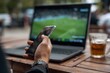 © LimeSky - Zoomed in image of a man s hand with a smartphone He s streaming a soccer match on his laptop and placing bets on his preferred team via an app