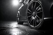 © Mariya - Close-up of shiny car wheels on wet asphalt with bright light source creating dramatic reflections in a dark environment