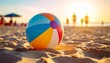 © rifqi - A colorful beach ball rests on sand with blurred figures and sunset.