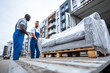 © Anastasiia Havelia - Two professional male international movers unloading furniture from a truck, one is wearing blue overalls and the other man has a white shirt. The sofa is covered by plastic wrap.