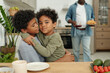 © pressmaster - Two young boys with curly hair hugging in cozy kitchen setting while African American male adult holding plate and glass is in background, creating a warm family atmosphere