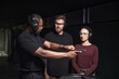 © DragonImages - African American man instructing Caucasian young adult man and woman at indoor shooting club, all wearing protective earmuffs and safety glasses, focusing on handgun handling