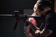 © DragonImages - Man wearing protective earmuffs instructing Caucasian young woman on holding rifle at shooting club indoor range, both focusing on firearm handling